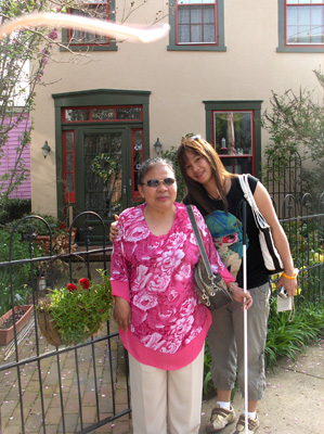 Nanta and Ann stand in front of a fence of a charmng yellow home with flowers on the fence.